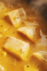 Close-up shot of the chicken simmering in the curry sauce in the pan, on the white marble cooktop. The sauce has thickened, and the chicken pieces are tender and cooked through