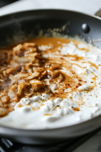 Close-up shot of the chicken simmering in the curry sauce in the pan, on the white marble cooktop. The sauce has thickened, and the chicken pieces are tender and cooked through