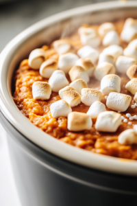 Close-up shot of mini marshmallows being sprinkled on top of the sweet potato casserole in the slow cooker. The marshmallows are gently melting on top as the casserole cooks for a few more minutes, all on the white marble cooktop