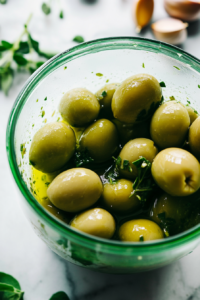 Close-up shot of Castelvetrano olives being poured into a food processor along with 6 tablespoons of brine. The fresh herbs and garlic are prepared on the side