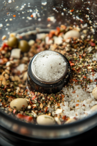 Close-up of a spoon sprinkling salt and pepper into the olive mixture while in the food processor, ensuring the flavors are perfectly balanced.