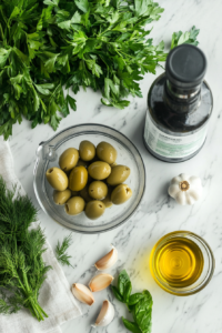 A neatly arranged countertop featuring 2 cups of Castelvetrano olives with brine, fresh parsley, dill, basil, garlic cloves, and a bottle of olive oil. Everything is set near a food processor, ready for blending.