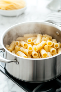 Close-up shot of a large stainless steel pot on the white marble cooktop, with salted water boiling vigorously. Gluten-free pasta is being added to the pot