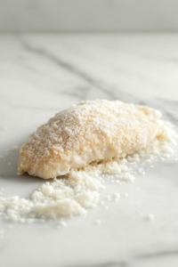 Close-up shot of a black skillet on the white marble cooktop, sizzling with ghee as breaded chicken cooks inside. The chicken is golden brown and crisp, with steam rising from the pan.
