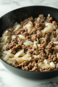 Ground beef (or a mix of beef and Italian sausage) being added to the softened onions and garlic in the Dutch oven. The meat is browning evenly as a wooden spoon breaks it apart.