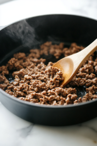 Close-up of a Dutch oven on a white marble cooktop, with ground beef or sausage being browned over medium heat. The meat is evenly cooked and ready to be drained