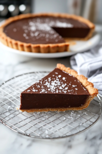The chilled chocolate tart sitting on a wire rack, with flaky sea salt sprinkled on top. A slice is served on a plate, ready to enjoy