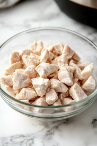Close-up shot of chopped chicken in a glass bowl on the white marble cooktop, being coated with almond flour. The chicken is tossed gently to ensure an even coating