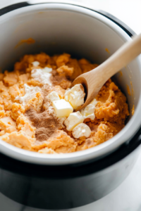 Close-up shot of mashed sweet potatoes being added to the slow cooker on the white marble cooktop. Melted butter, heavy cream, eggs, brown sugar, and cinnamon are stirred into the mixture with a wooden spoon.
