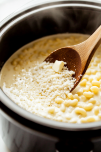 Close-up shot of uncooked elbow macaroni being added to the slow cooker with melted butter, evaporated milk, whole milk, dry mustard, and salt. The mixture is being stirred together with a wooden spoon over the white marble cooktop
