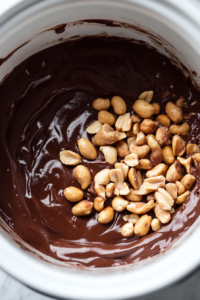 Close-up shot of the melted chocolate mixture in the slow cooker, with salted dry roasted peanuts, unsalted dry roasted peanuts, and cocktail peanuts being stirred into the chocolate blend. The slow cooker sits on the white marble cooktop