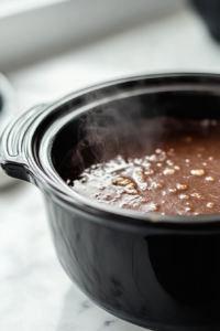 Close-up shot of a slow cooker on the white marble cooktop, set to Low heat and cooking the chocolate mixture. The lid is on, and condensation forms on the edges, indicating the gentle cooking process.
