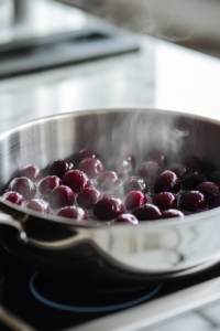 Close-up shot of a shiny stainless steel saucepan on the white marble cooktop, with frozen cherries simmering to release their juices. Steam rises from the pan, and the cherries glisten.