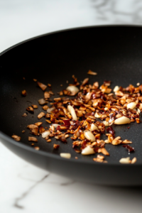 Close-up shot of minced garlic and ginger sizzling in the same black non-stick skillet on the white marble cooktop. The mixture is fragrant and browning slightly