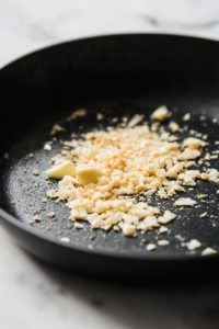 The same black skillet on the white marble cooktop, with the remaining butter melting and minced garlic being sautéed. The butter is sizzling, and the garlic is golden and aromatic
