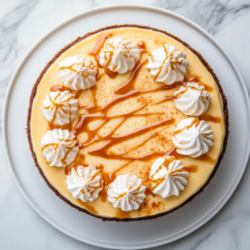 A creamy cheesecake on a plate, drizzled with salted butterscotch sauce and topped with whipped cream. A fork is beside the plate, ready to dig in.
