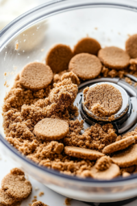 Close-up shot of vegan ginger cookies being crushed into crumbs in a food processor. The cookies are broken down into small pieces, ready to be mixed with melted vegan butter for the crust.