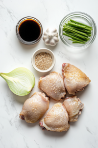Top-down shot of the ingredients on a kitchen counter: chicken thighs with skin on, a sliced onion, tamari sauce in a small bowl, a glass of water, minced garlic in a small dish, salt, pepper, green onion, and sesame seeds. All items are arranged neatly with a rustic wooden backdrop