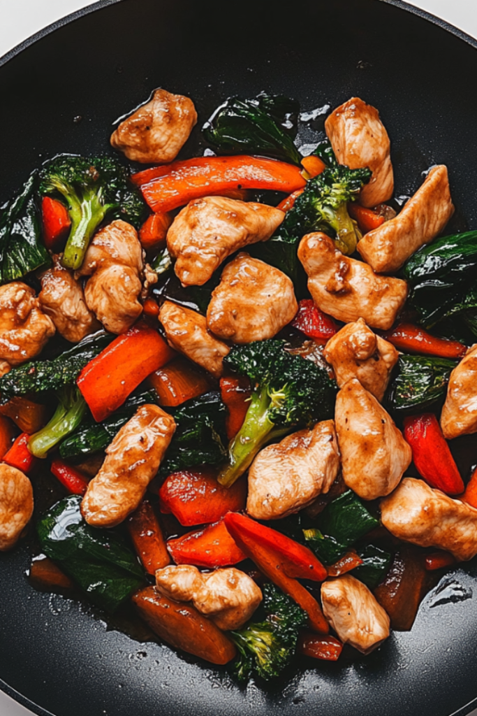 Close-up shot of a spoon serving the stir fry onto a plate from the black non-stick skillet on the white marble cooktop. The chicken and vegetables are evenly coated with the sauce, ready to enjoy