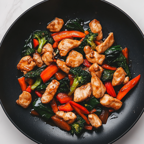 Close-up shot of a spoon serving the stir fry onto a plate from the black non-stick skillet on the white marble cooktop. The chicken and vegetables are evenly coated with the sauce, ready to enjoy