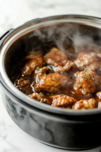 Overhead shot of raw chicken thighs layered at the bottom of a slow cooker, topped with sliced onions and minced garlic, with tamari sauce and water being poured over the ingredients.