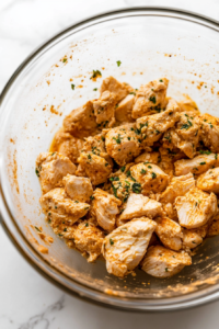 Close-up shot of diced chicken breasts being mixed with plain yogurt, minced garlic, grated ginger, garam masala, cumin, paprika, salt, and a splash of lemon juice in a glass bowl. The bowl sits on the white marble cooktop, ready to be covered and refrigerated.
