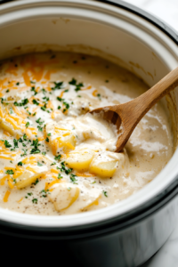 Close-up shot of a wooden spoon stirring the creamy mixture of potatoes, sour cream, cheese, and soup in the slow cooker on the white marble cooktop. The ingredients are blending into a rich, cheesy mixture.
