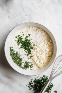 A large mixing bowl on a white marble cooktop containing almond flour, Savory Chicken Soup Keto Chow, sage, parsley, and baking powder. A whisk rests beside the bowl