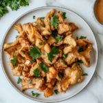 A serving plate of golden-fried oyster mushrooms arranged neatly, garnished with fresh parsley. A small bowl of dipping sauce sits beside the plate on a rustic table setting with a napkin, fork, and a glass of lemonade.