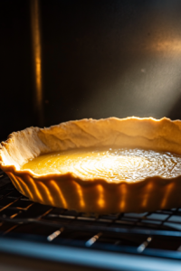 The tart crust lined with parchment paper, filled with baking beads, and placed into the oven at 350°F (175°C). The crust is lightly golden.