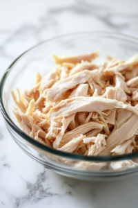 Close-up of shredded chicken breast in a clear glass bowl on a white marble cooktop, ready for use in the soup. The chicken is tender, with juicy fibers, and the bowl is pristine, reflecting the light.