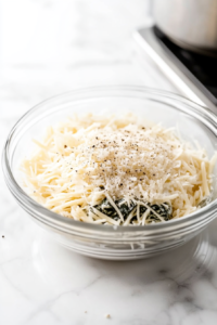 Close-up of a clear glass bowl on a white marble cooktop, filled with softened cream cheese, chopped raw spinach, minced garlic, grated Parmesan, mozzarella, ground nutmeg, black pepper, and kosher salt. A spoon is blending the creamy, colorful filling.