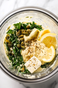 A blender with ingredients for the vegan Caesar dressing: vegan mayo, Dijon mustard, capers, nutritional yeast, lemon juice, garlic powder, salt, and pepper, being blended until smooth and creamy.