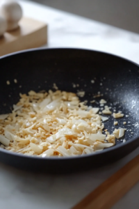 Close-up shot of the black skillet on the white marble cooktop, with minced garlic and sliced cremini mushrooms added to the golden onions, cooking and softening.