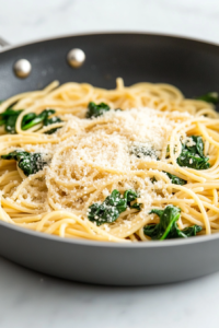 Close-up shot of the skillet on the white marble cooktop, with a sprinkle of vegan parmesan cheese on top of the pasta and spinach mixture, ready to serve