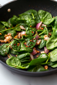 Close-up shot of fresh spinach leaves being added to the sautéed shallots, garlic, and red pepper flakes in the black skillet on the white marble cooktop. The spinach starts to wilt.