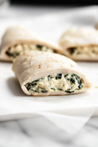 Close-up of stuffed chicken rolls, seam-side down, on a baking sheet lined with parchment paper, resting on the white marble cooktop. The rolls are ready to chill.