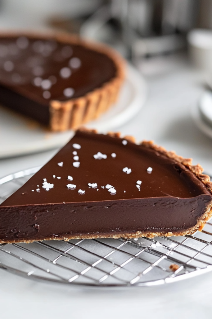 The chilled chocolate tart sitting on a wire rack, with flaky sea salt sprinkled on top. A slice is served on a plate, ready to enjoy