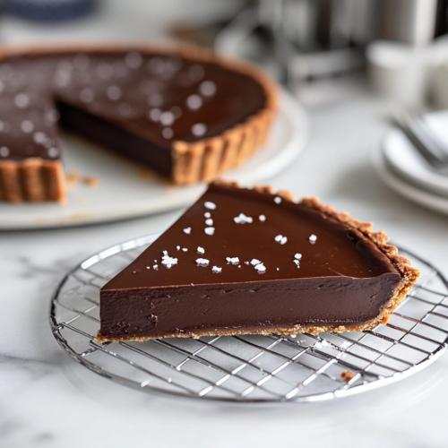 The chilled chocolate tart sitting on a wire rack, with flaky sea salt sprinkled on top. A slice is served on a plate, ready to enjoy