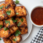 Plate of crispy fried tofu 'chicken' arranged in a stack, garnished with fresh parsley. A small bowl of dipping sauce is on the side, and a checkered napkin is folded next to the plate on a rustic table setting.