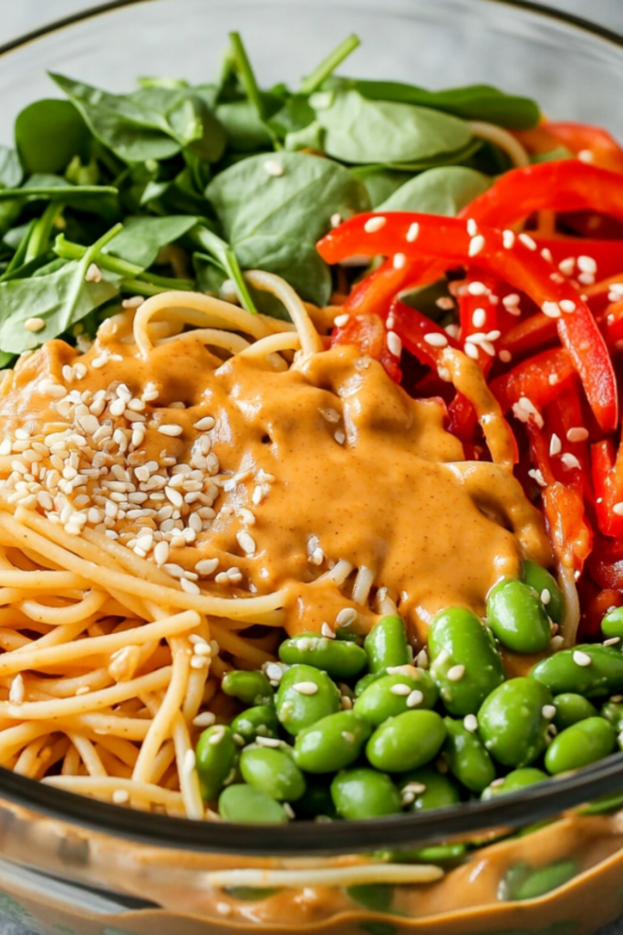 Using tongs to toss the ingredients in a large bowl, mixing the pasta and vegetables with the creamy peanut sauce. The dish is garnished with sesame seeds and ready to serve.