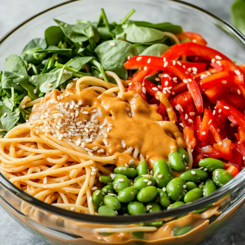 Using tongs to toss the ingredients in a large bowl, mixing the pasta and vegetables with the creamy peanut sauce. The dish is garnished with sesame seeds and ready to serve.