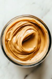 A close-up of a hand whisking together peanut butter, sriracha, soy sauce, rice vinegar, water, maple syrup, toasted sesame oil, garlic powder, and ginger powder in a mixing bowl. The mixture is becoming smooth and creamy