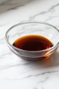 Close-up shot of a glass bowl on the white marble cooktop, filled with stir fry sauce, sesame oil, and chili sauce being whisked together until smooth and well combined.