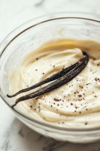A close-up shot of a glass mixing bowl on the white marble cooktop, with a vanilla bean pod being scraped to release the seeds into the creamy mixture. The seeds swirl in as they are blended.