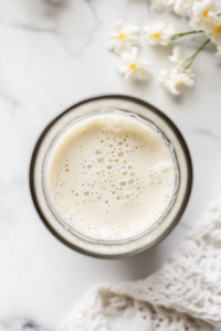 Close-up shot of a smooth glass blender on the white marble cooktop, containing silken tofu being blended until creamy. The blender lid is slightly lifted to reveal the thick, smooth texture of the tofu inside.