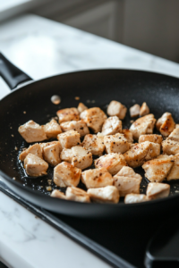 Chicken chunks sizzling in a black skillet on a white marble cooktop, being sprinkled with garlic, basil, salt, pepper, and oregano. A wooden spatula is stirring the chicken as it browns