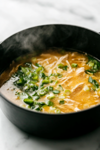 Close-up of a black pot on a white marble cooktop, deglazing with chicken stock while adding coconut milk, shredded chicken, lime juice and zest, fish sauce, lemongrass, and scallions. The mixture gently simmers, with steam rising.