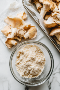 Overhead view of a breading station with a bowl of seasoned flour and cornstarch mixture next to a bowl of marinated oyster mushrooms. A few mushrooms are being coated while others rest breaded on a parchment-lined tray.