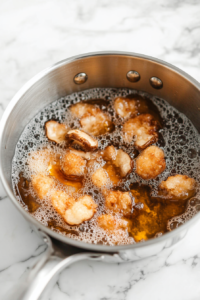 Action shot of oyster mushrooms frying in a saucepan filled with bubbling vegetable oil. The mushrooms are golden brown, and a slotted spoon is in the foreground, ready to lift a piece